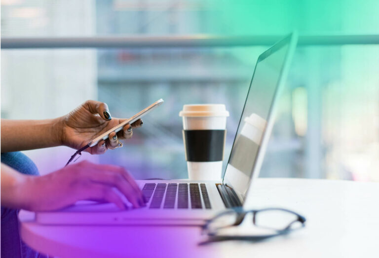 Person som sitter vid datorn med en mobil i handen och takeaway-kaffe bredvid